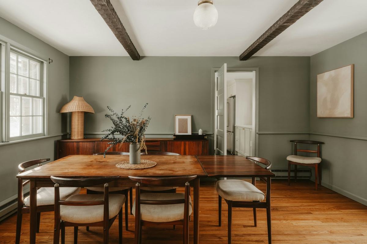 Rustic dining room with a solid wood table, upholstered chairs, exposed ceiling beams and a sideboard against a sage green wall